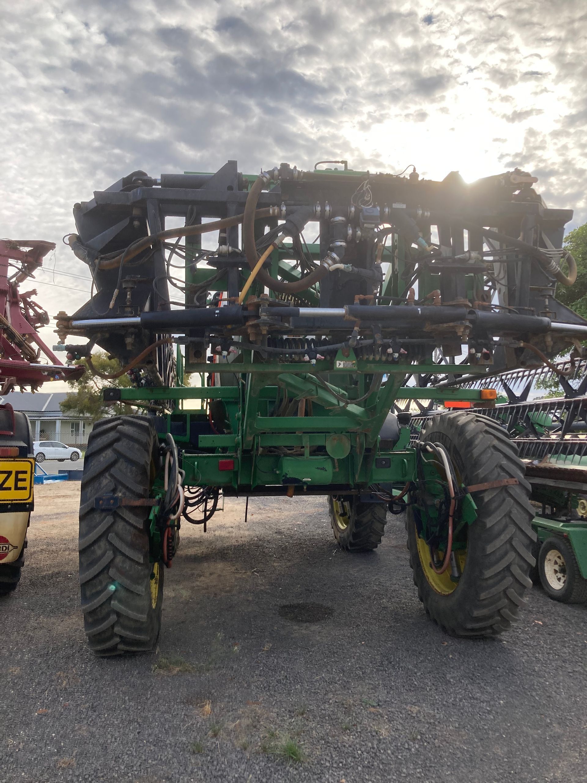 Large green agricultural sprayer with wide boom arms parked on gravel at sunset