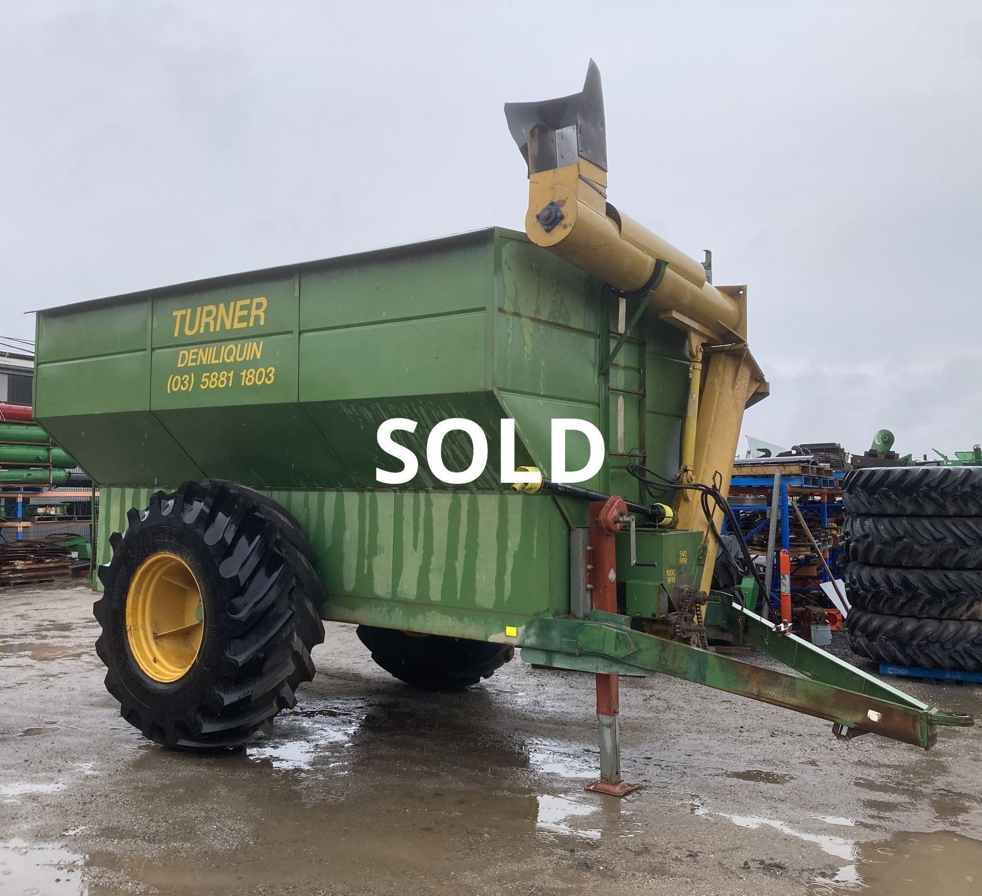 Green farm trailer with large tires and “SOLD” painted on the side in a muddy lot