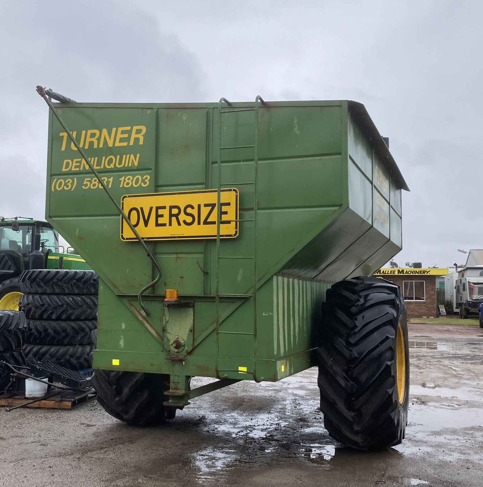 Green oversized farm trailer with yellow warning sign, parked on wet pavement in a farmyard