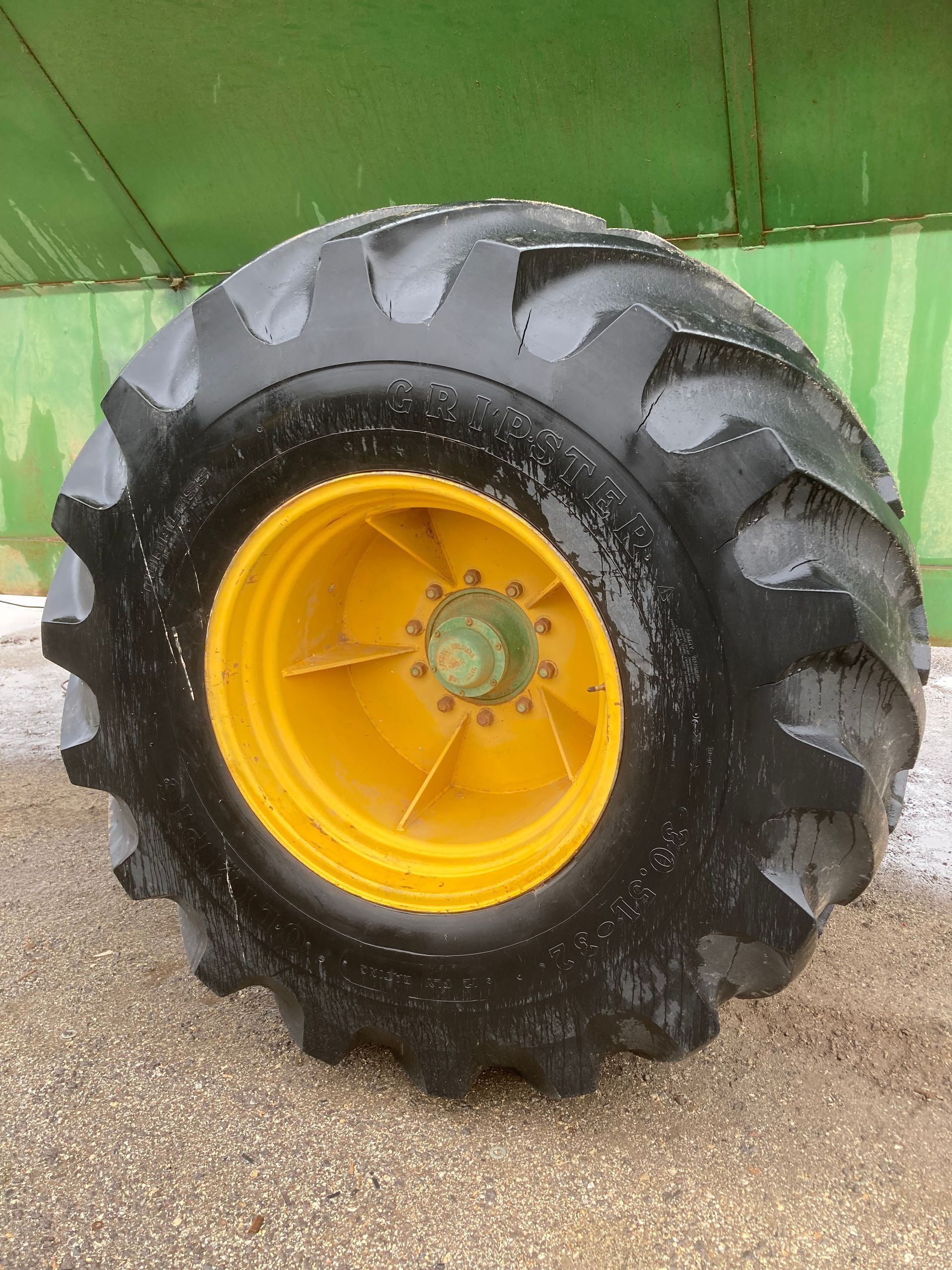 Large black tractor tire with yellow rim beside a green vehicle, wet pavement beneath it