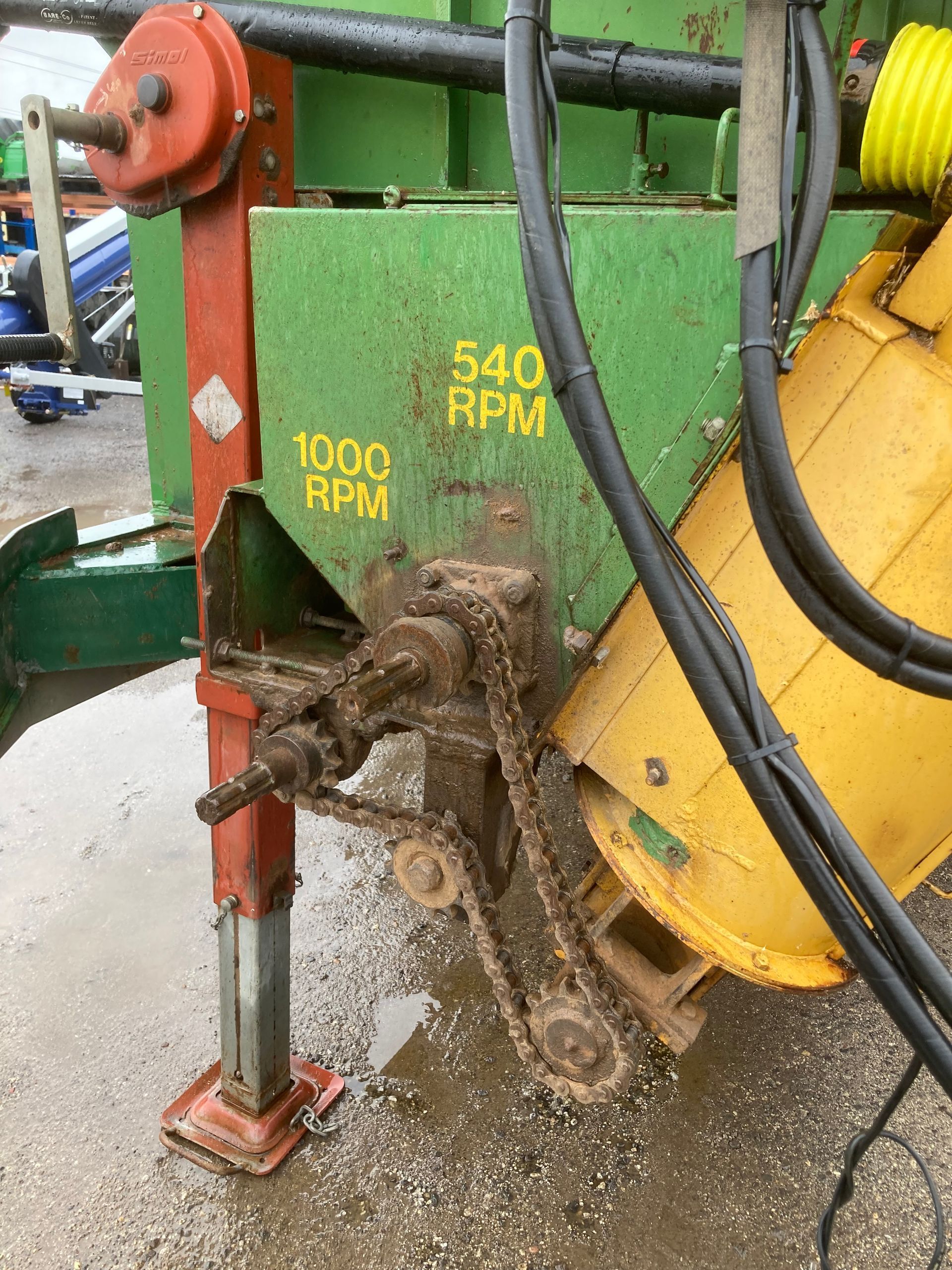 Green agricultural machine with yellow drum and muddy chain, labeled 100 RPM and 540 RPM