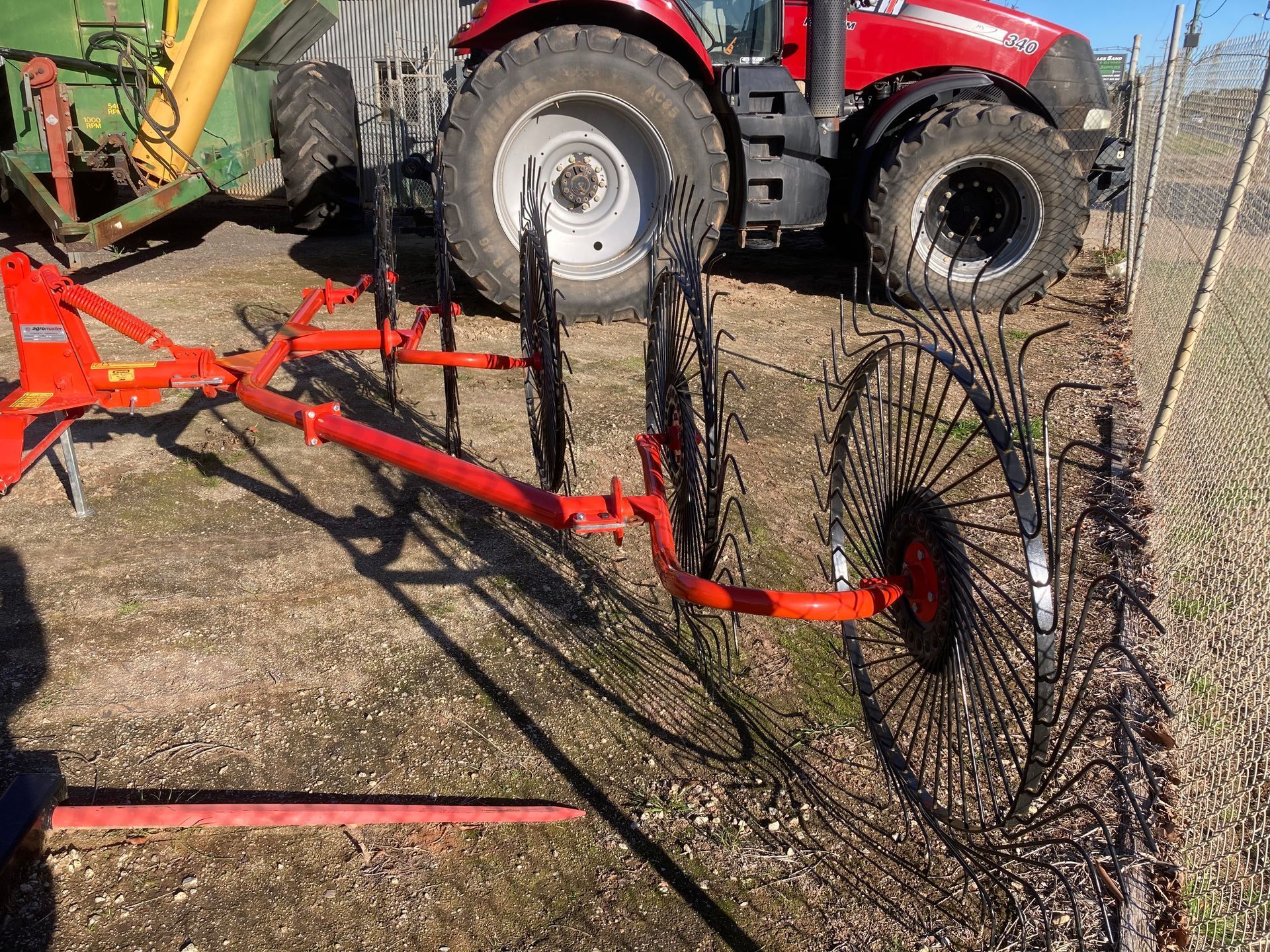 Red farm machine with large wheels and a metal reel in a gravel yard beside a tractor