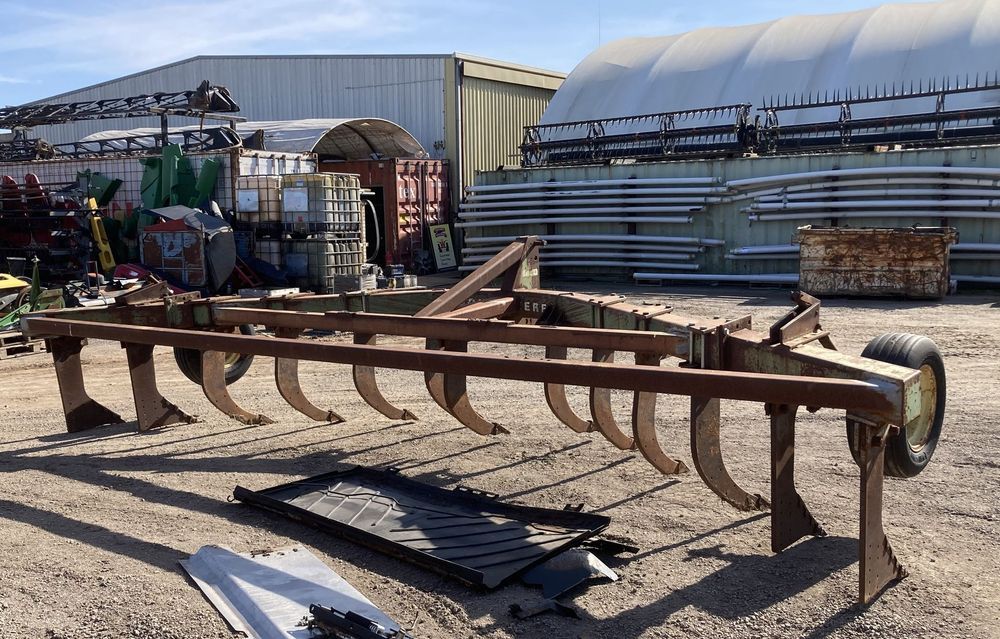 Rusty farm implement in a yard, with curved tines and metal sheds in the background