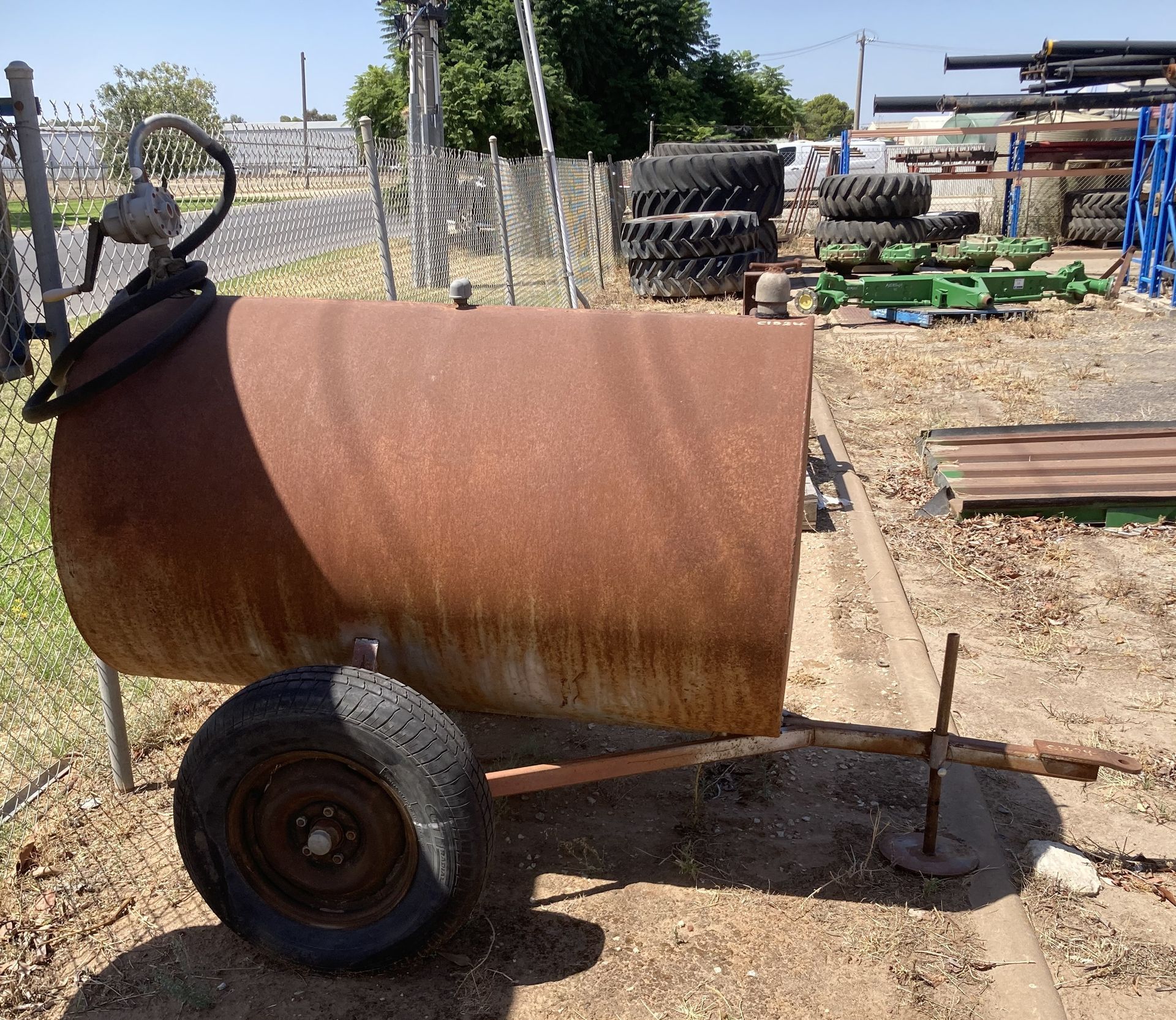 Rusty metal tank trailer in a dirt yard beside a fence and stacked equipment.