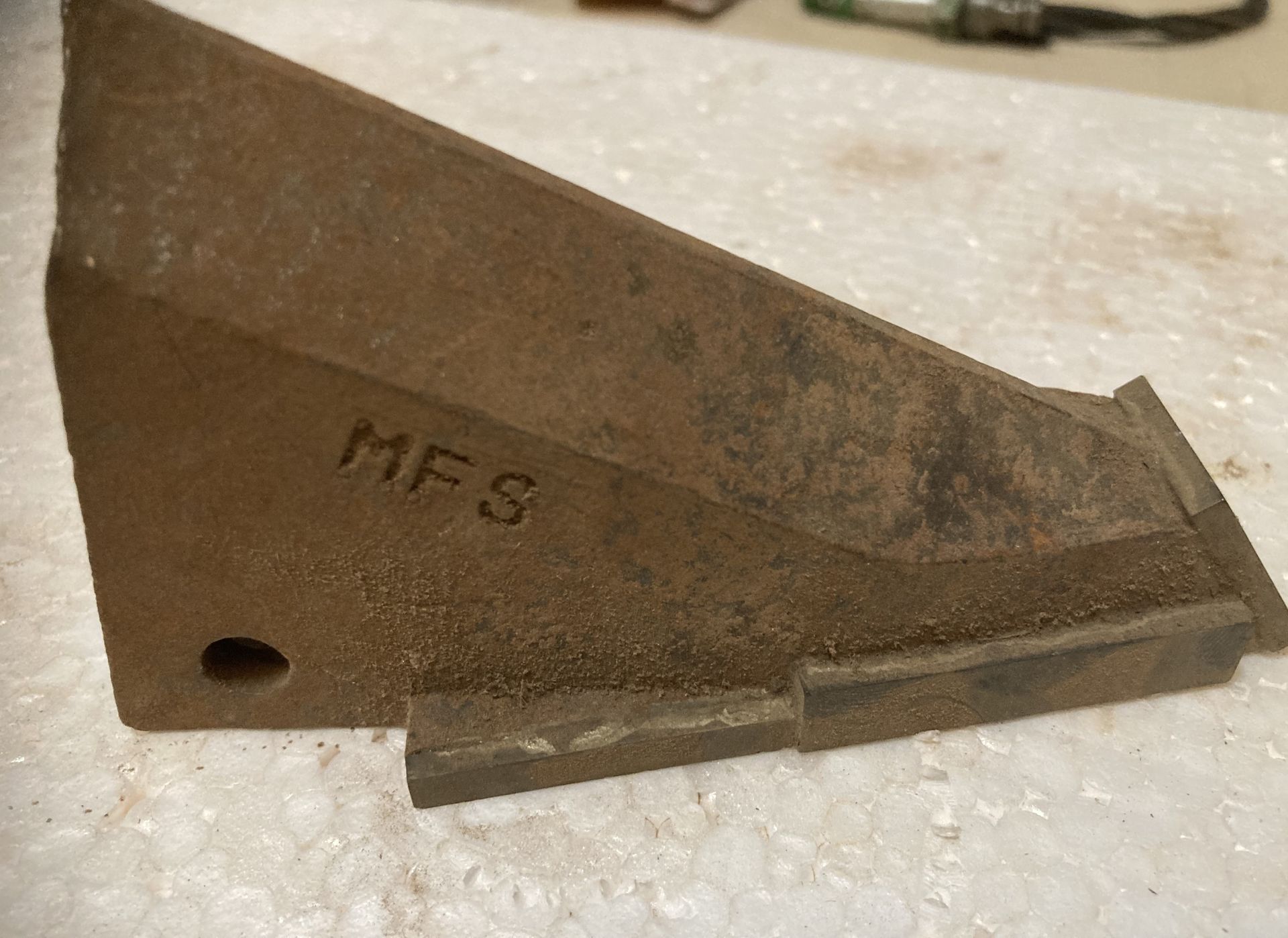 Rusty metal machine part with “MFS” stamped on it, resting on a light surface.