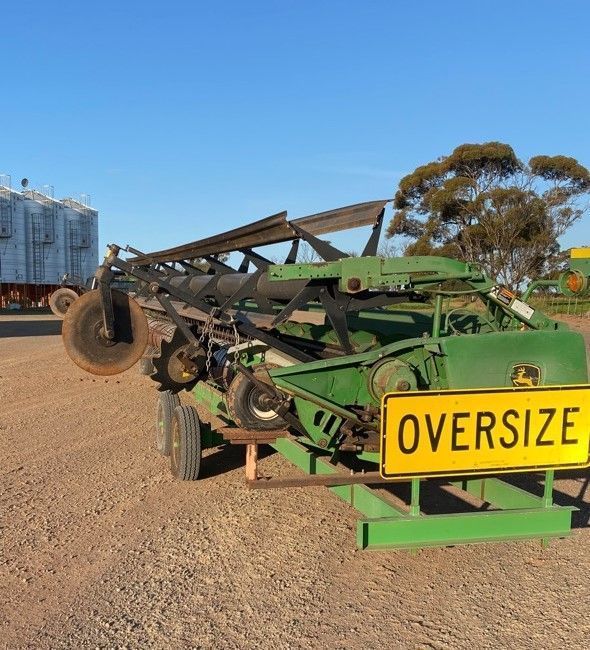 Green farm implement with yellow “OVERSIZE” sign parked on gravel lot under blue sky