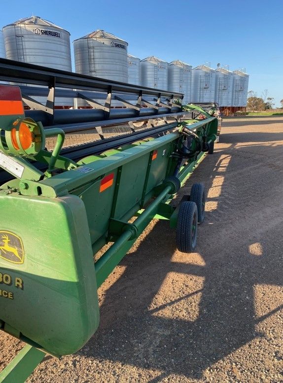 Green agricultural combine harvester parked on gravel beside silver grain silos under a blue sky
