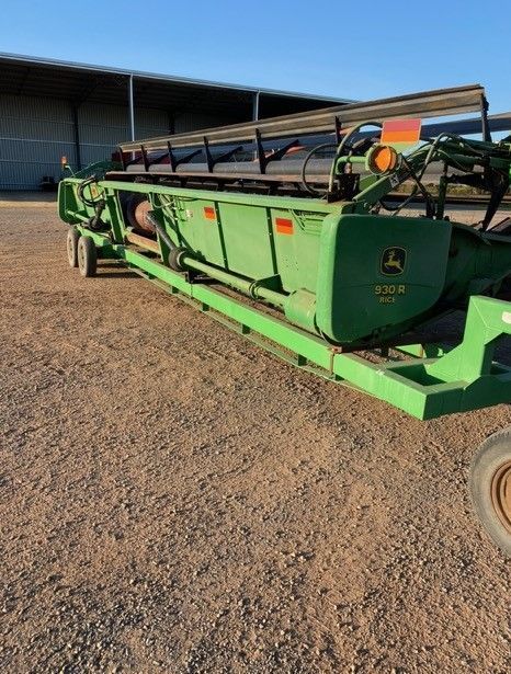 Green farm equipment trailer on gravel beside a barn under a clear blue sky