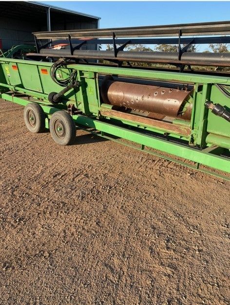 Green agricultural machine with a large brown drum on a gravel lot, beside a barn.