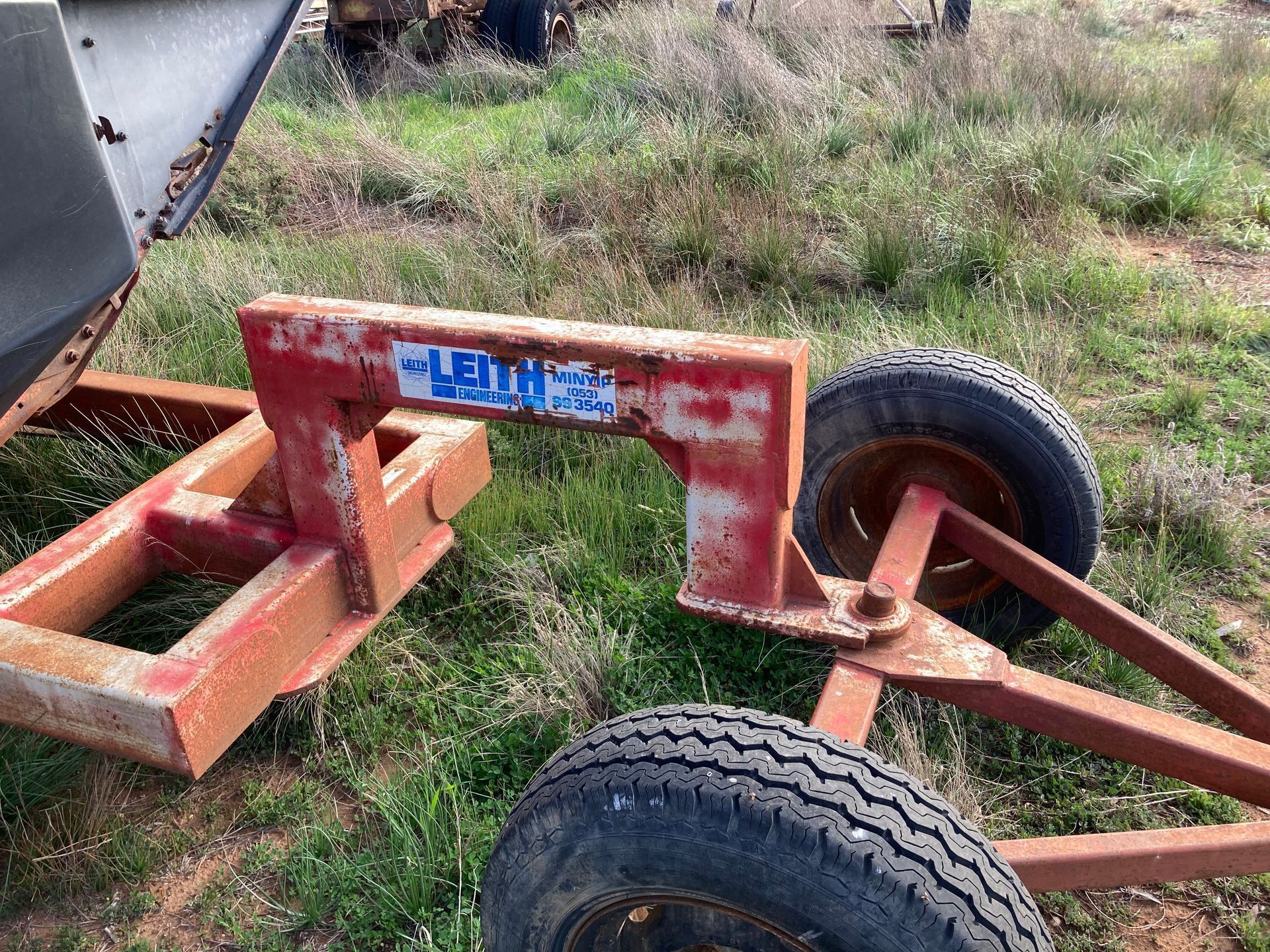 Rusty red trailer frame with two tires sitting in grass, showing a blue label on the center beam