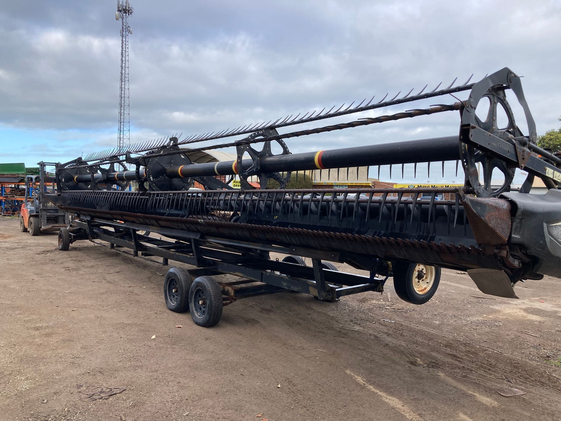 Large black agricultural combine header on a trailer in a dirt lot, with workers in the background