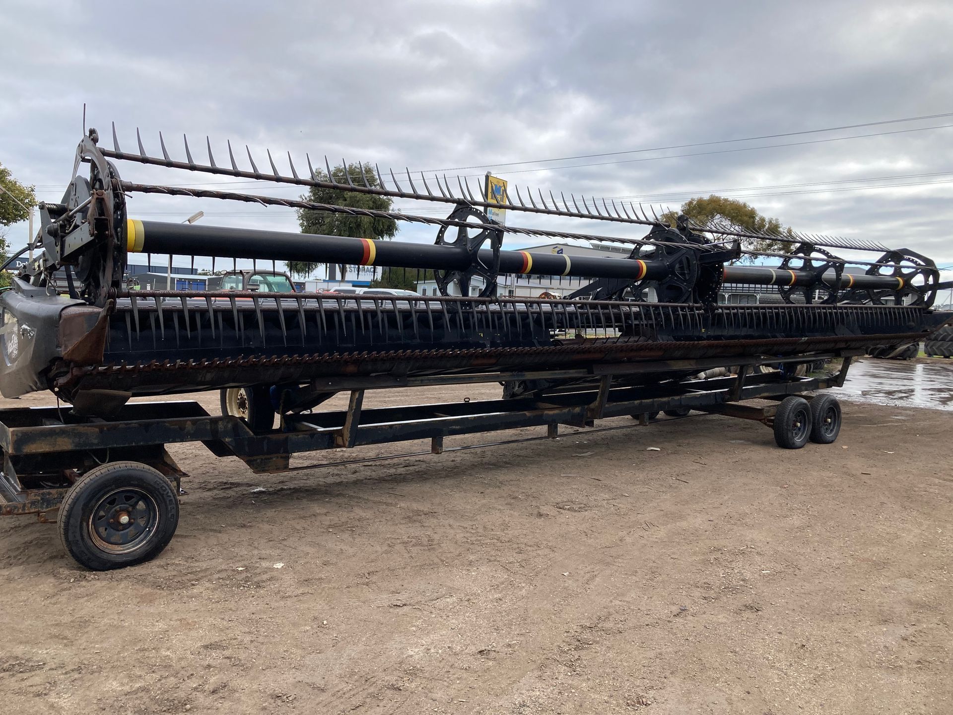 Large agricultural combine header on a trailer in a gravel lot under cloudy sky