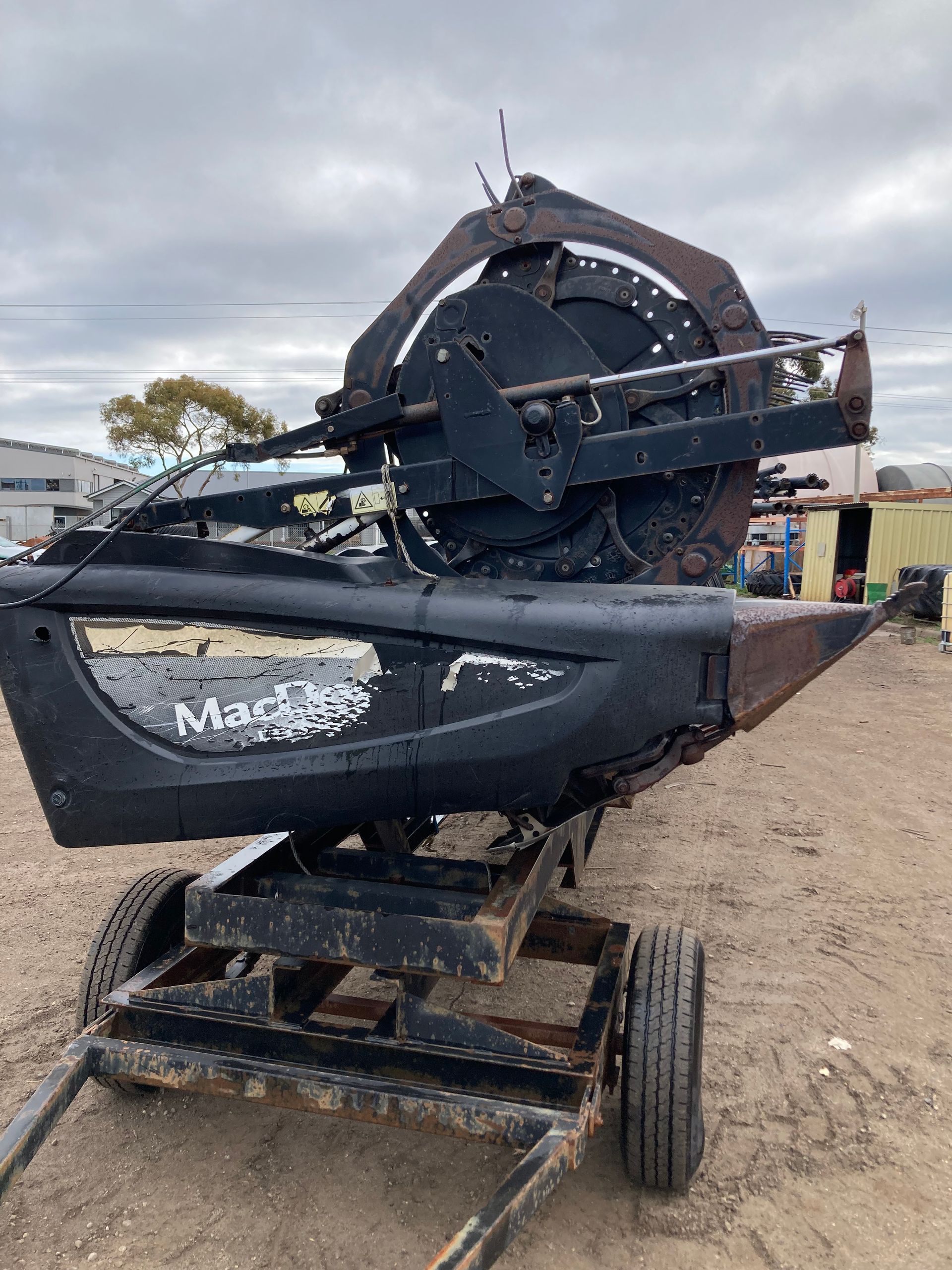 Damaged black MacDon farm machine with a broken reel, sitting on a gravel lot under cloudy skies