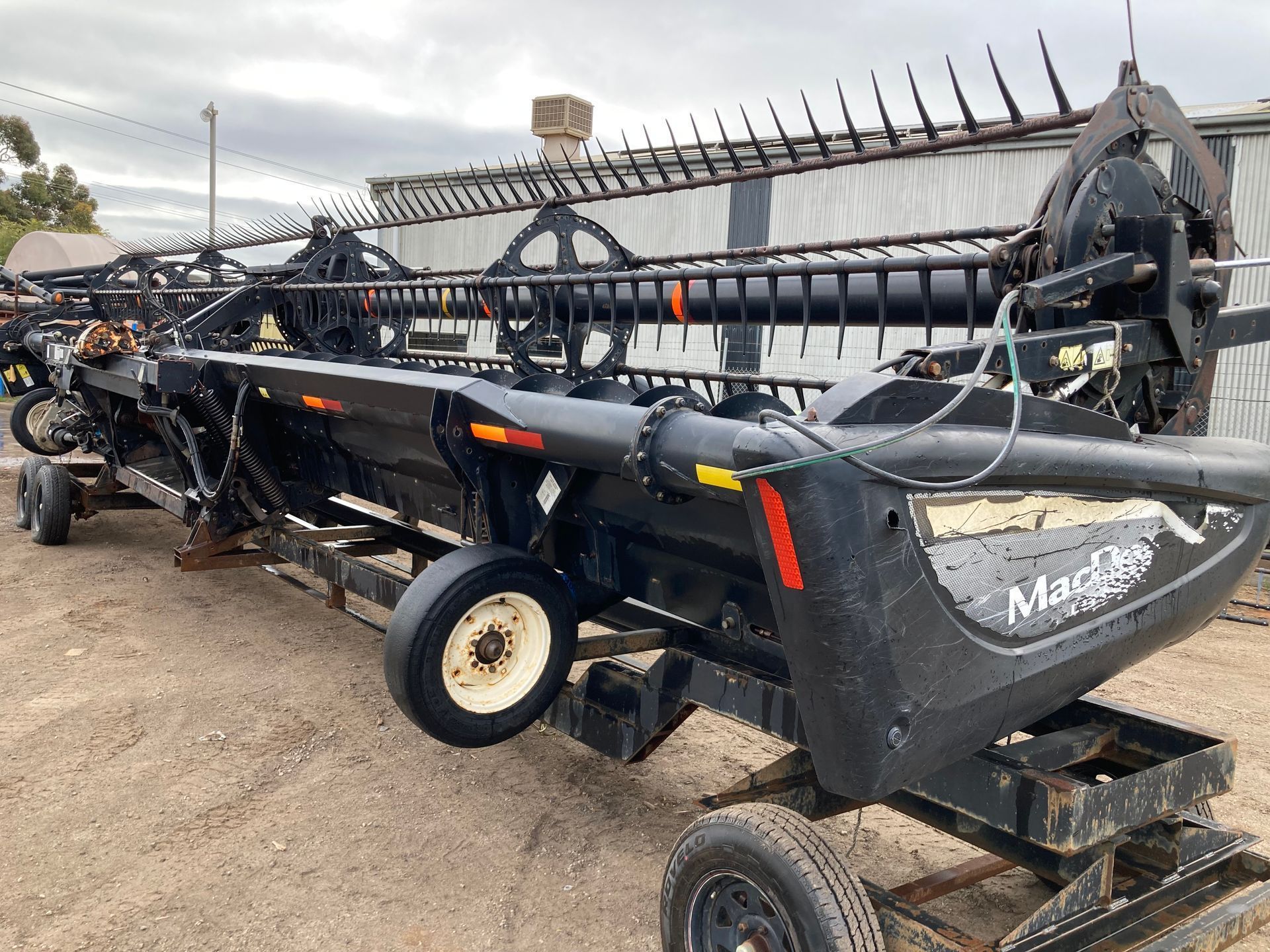 Black farm harvester header on a trailer in a muddy lot, with a long reel and metal frame