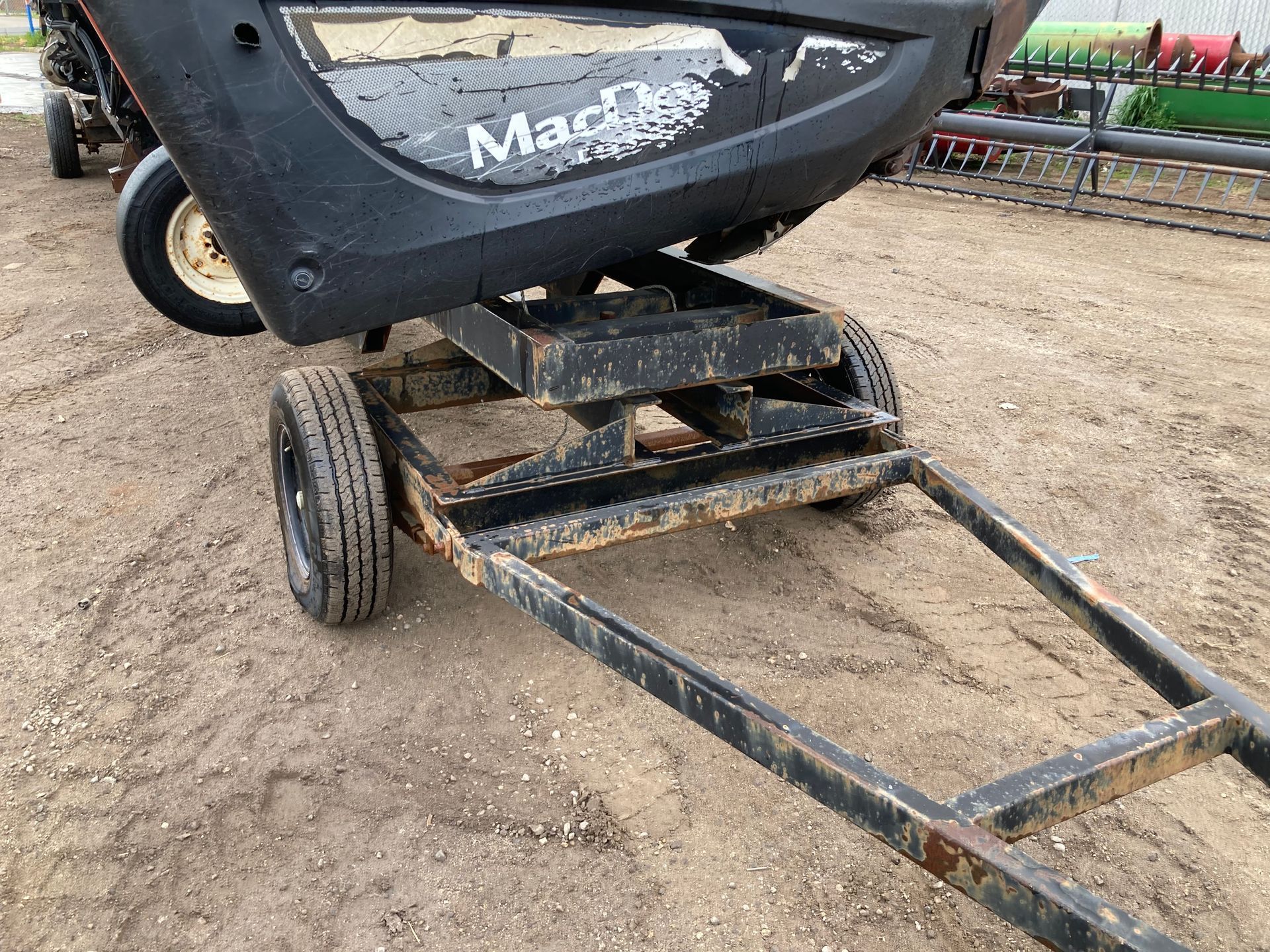 Black MacDon farm trailer hitch with metal frame and wheel on a dirt surface.