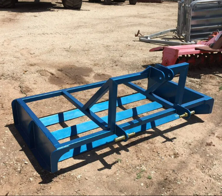 Blue metal skid-steer attachment on dirt lot, with black braces and red hydraulic mount.