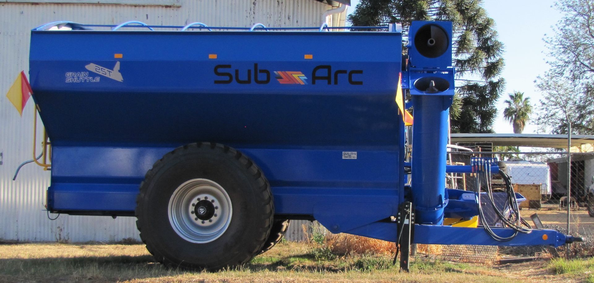 Blue agricultural machine on a trailer, labeled SubArc, parked outdoors on gravel near trees.