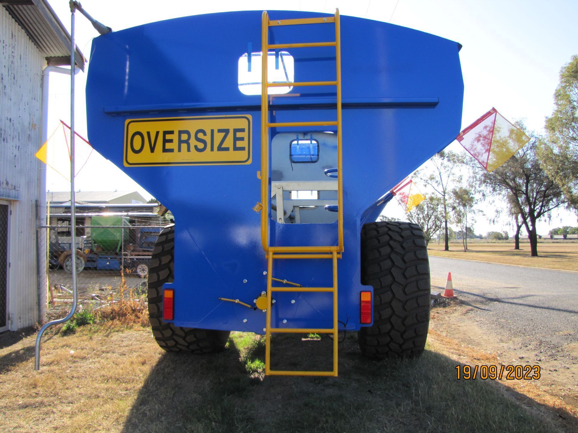 Rear view of blue oversized farm equipment with yellow ladder and oversized sign on a gravel road