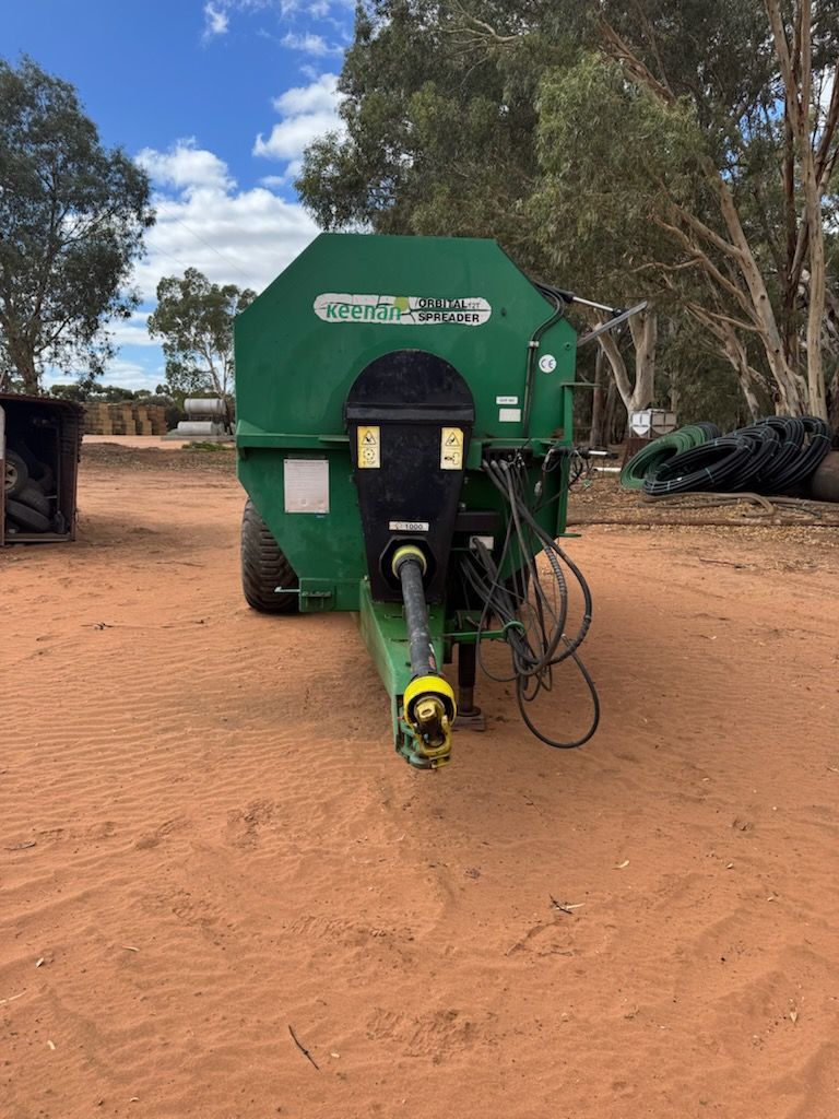 Green agricultural machine on a dirt farm track with trees and stacked tires in the background
