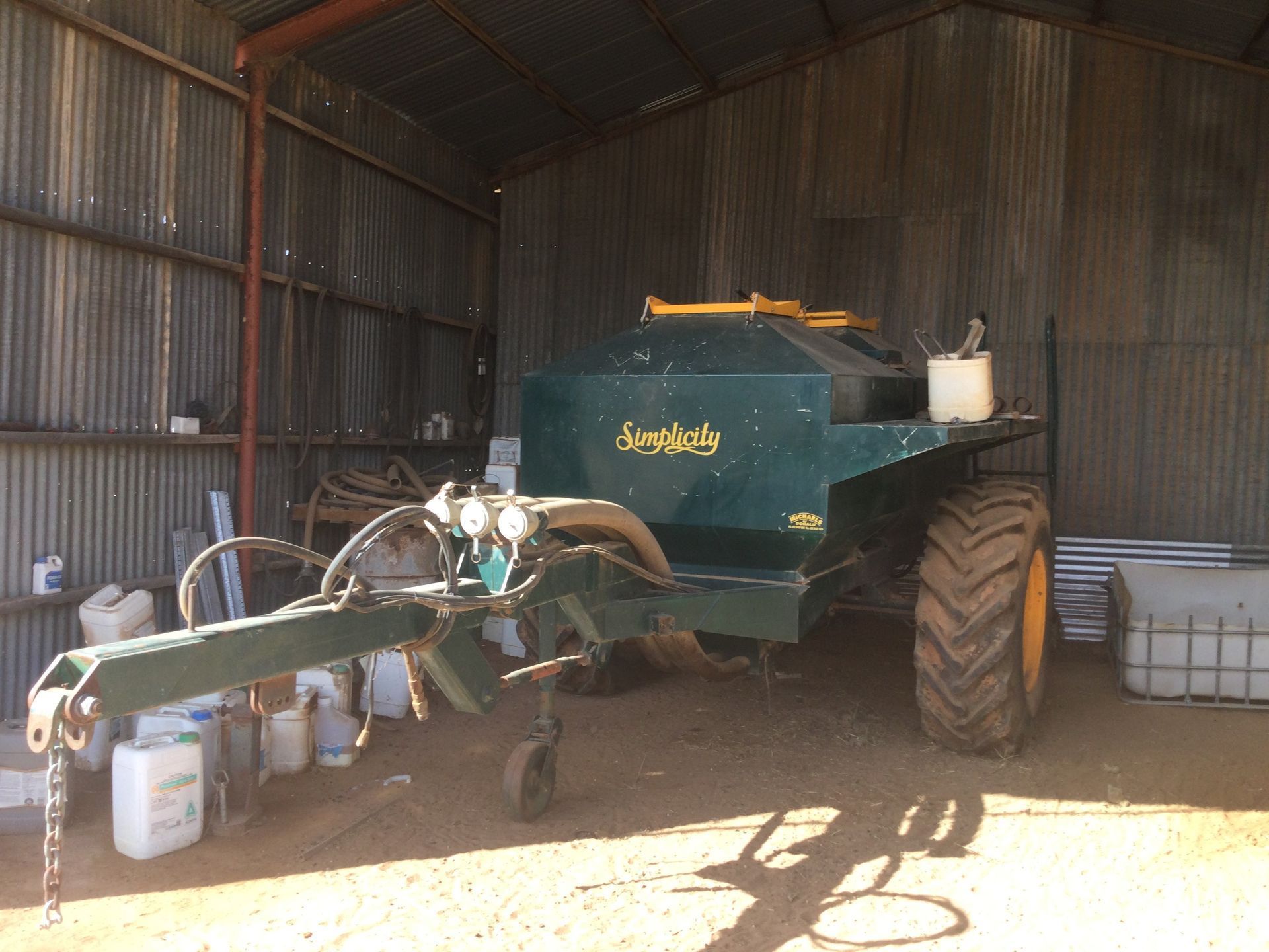 Green farm sprayer parked inside a metal shed, with hoses and white containers nearby.