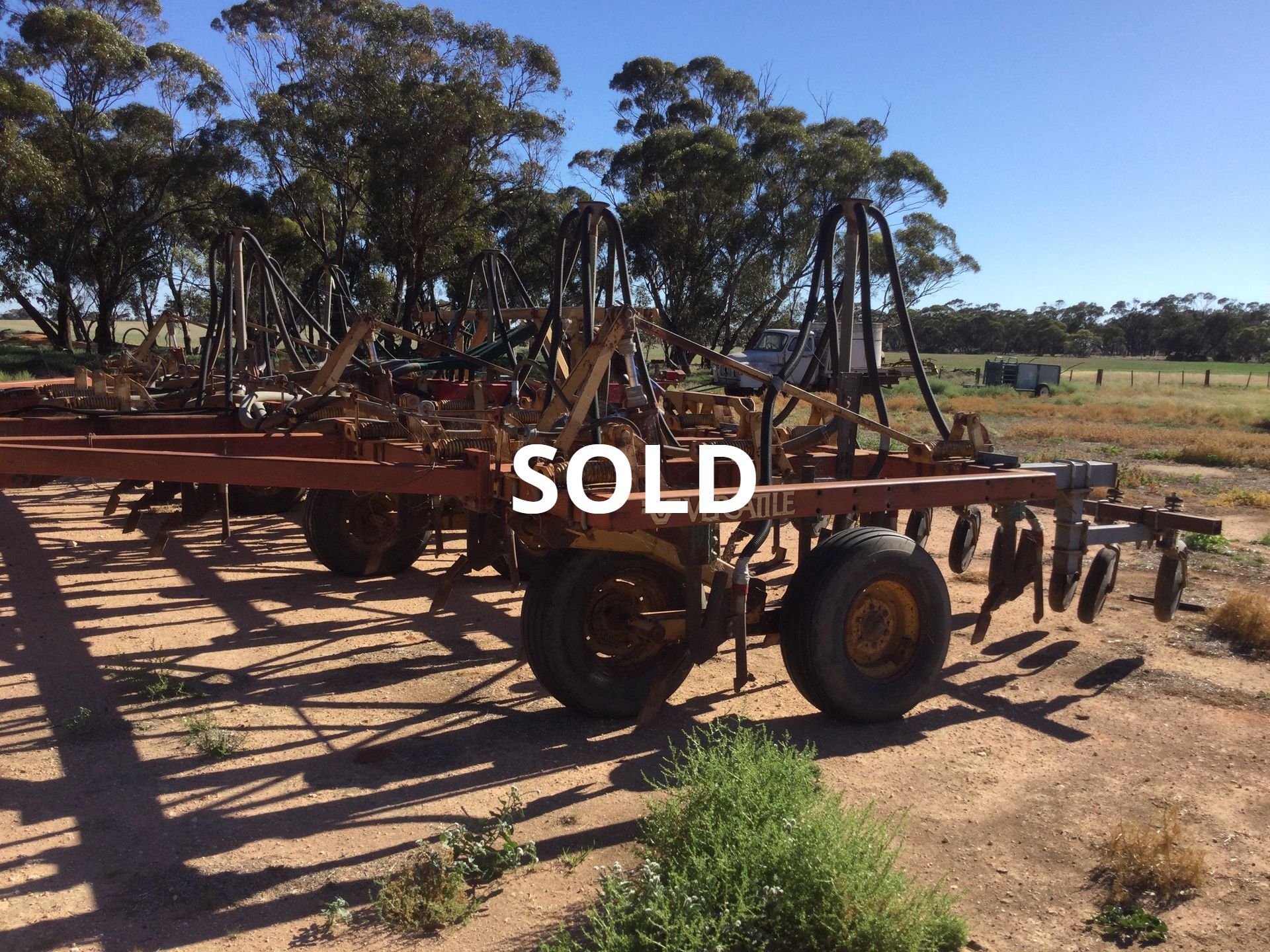 Old farm machinery on a dirt lot with a white SOLD sign, trees and blue sky in the background