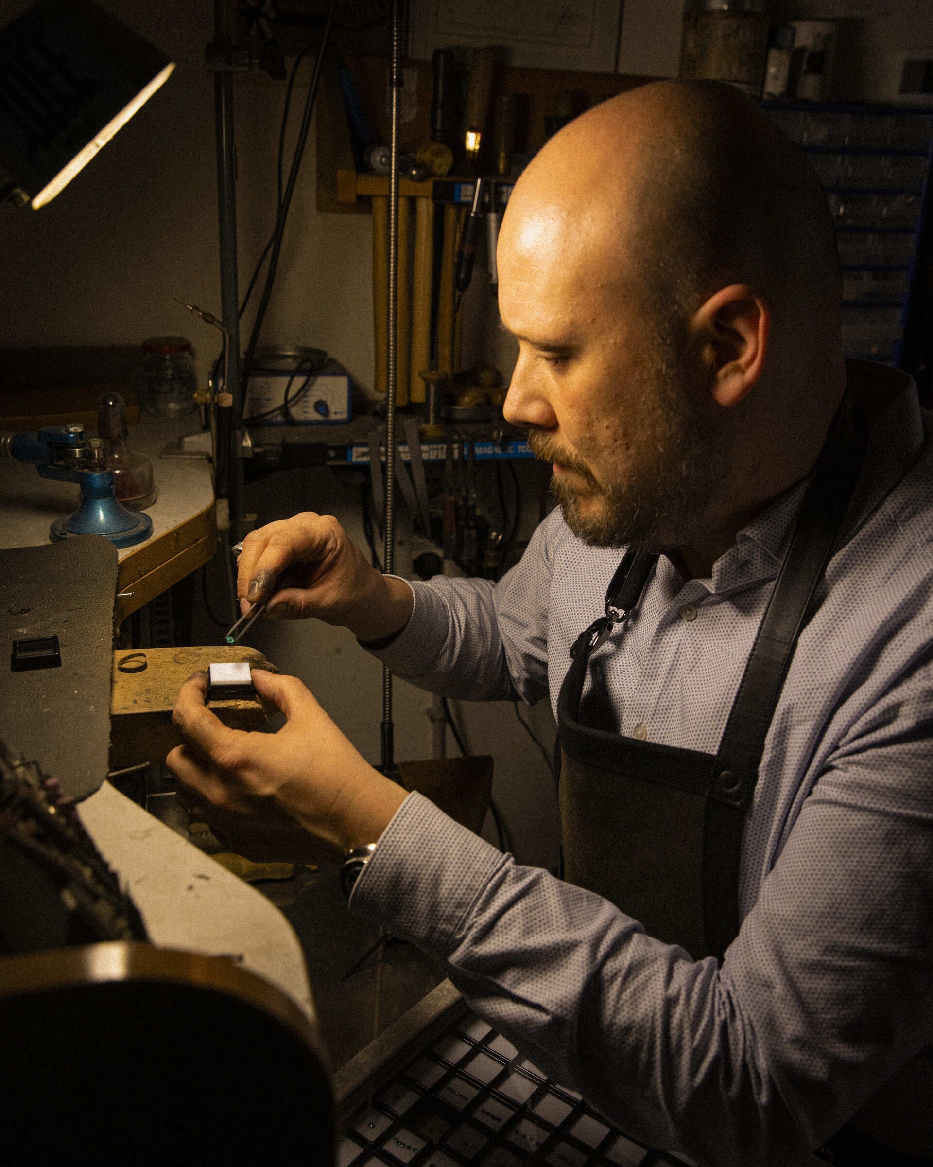 Andrea Carbone concentrato mentre lavora in un laboratorio orafo, posiziona una gemma su un gioiello utilizzando una pinzetta, illuminato da una lampada da tavolo Andrea Carbone concentrato mentre lavora in un laboratorio orafo, posiziona una gemma su un gioiello utilizzando una pinzetta, illuminato da una lampada da tavolo