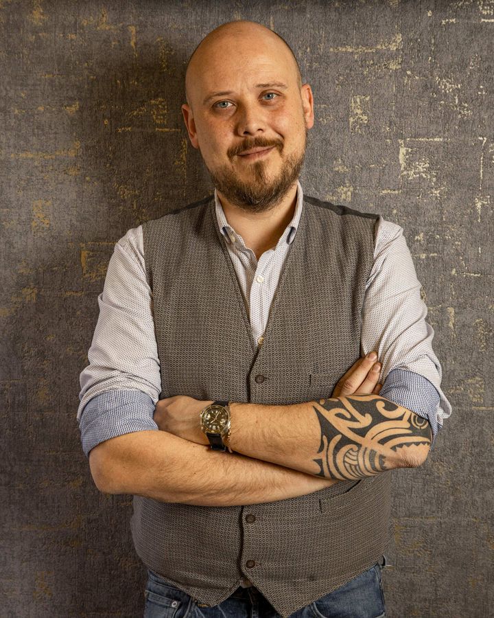 Andrea Carbone sorridente con tatuaggio tribale sull'avambraccio sinistro, indossa una camicia chiara e un gilet grigio, posando con le braccia incrociate su uno sfondo testurizzato