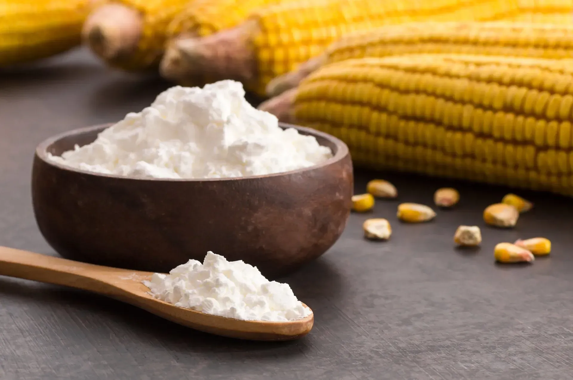 Bulk corn starch powder in a wooden bowl beside a wooden spoon filled with cornstarch.