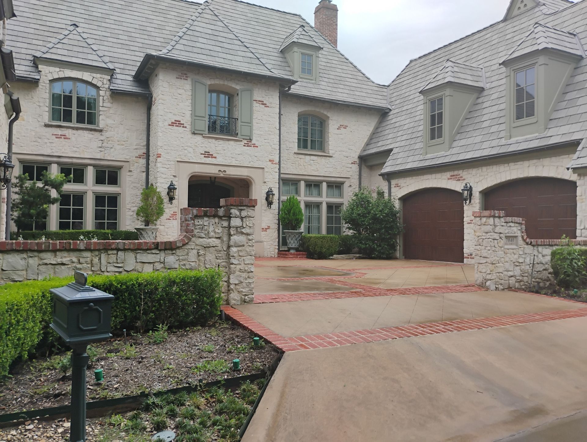 Stone mansion with garage and courtyard driveway, landscaped hedges, and slate roof under overcast light