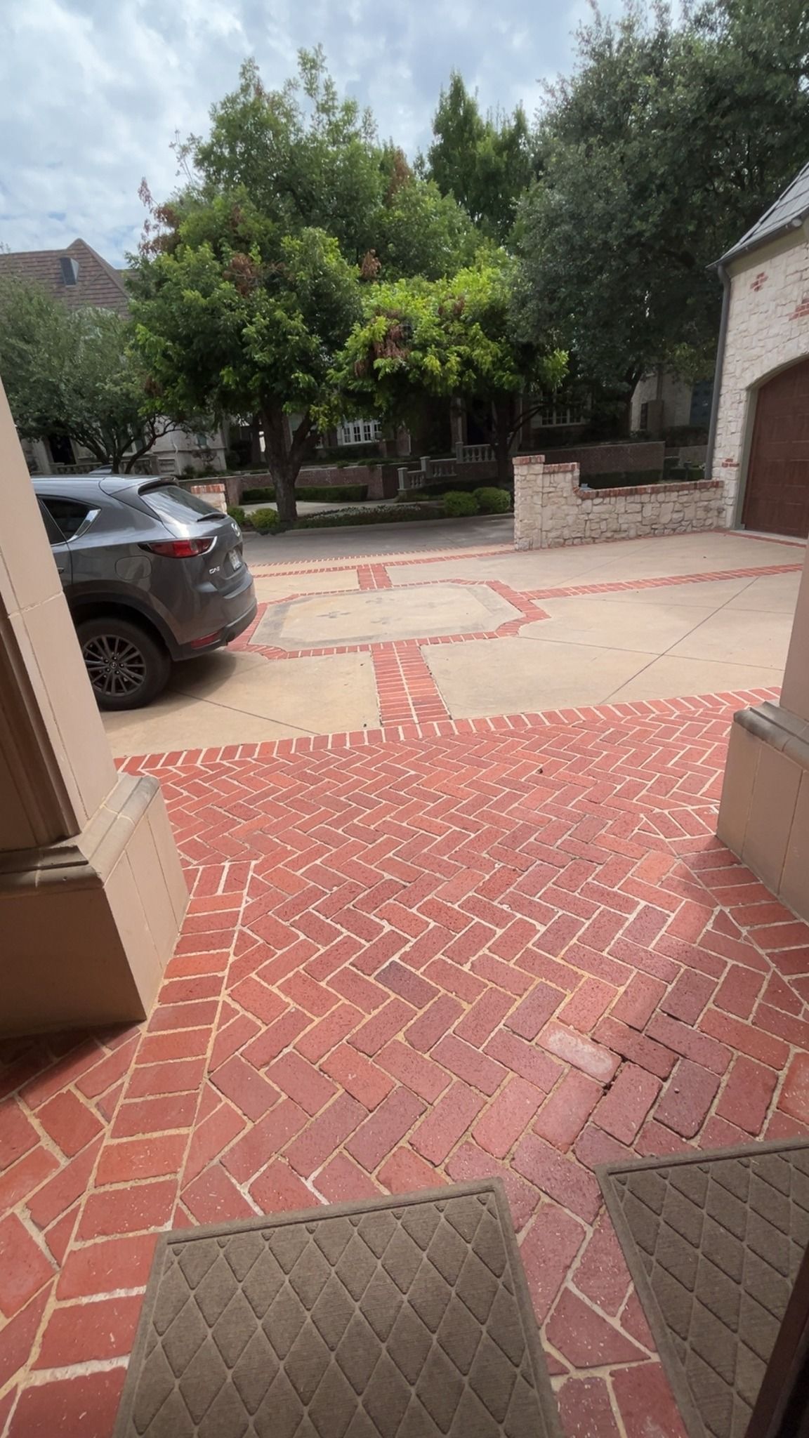 Brick-paved courtyard with a parked gray car, trees, and a stone bench in the background