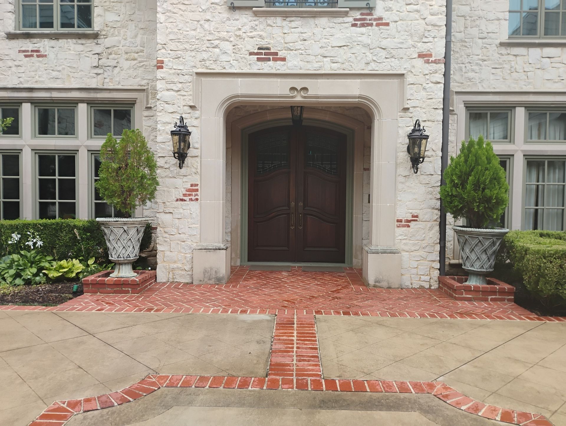 Stone house entrance with dark wooden double doors, red brick walkway, and potted topiary plants