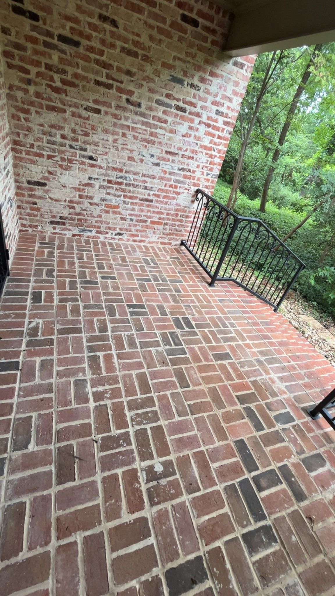 Brick porch with a sloped floor, brick wall, black railing, and trees beyond the edge