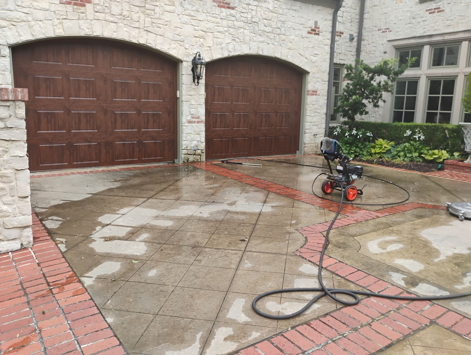 Pressure washing driveway in front of a stone house with garage doors and wet pavement