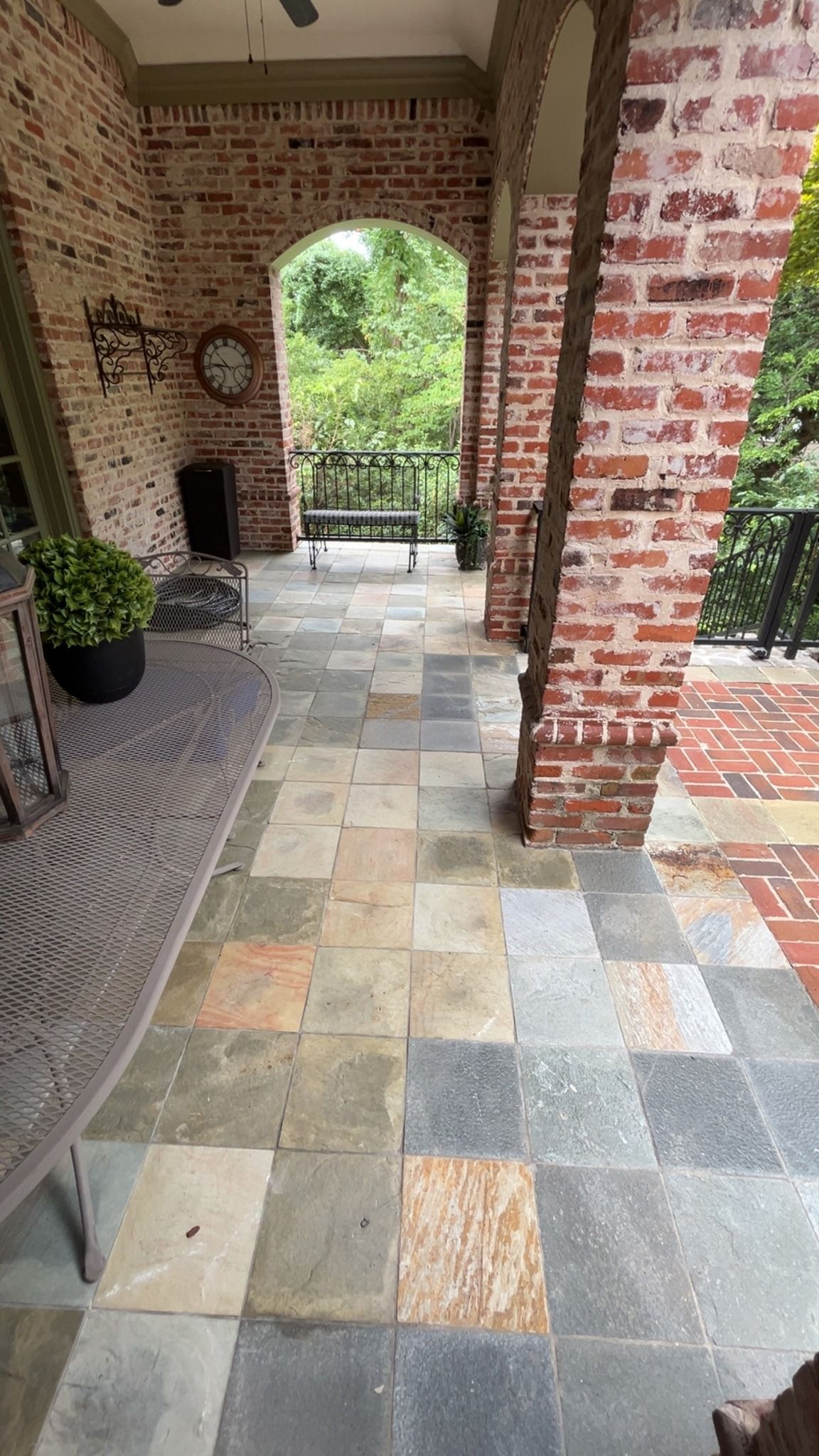 Brick porch with arched opening, patterned tile floor, and potted plants overlooking greenery