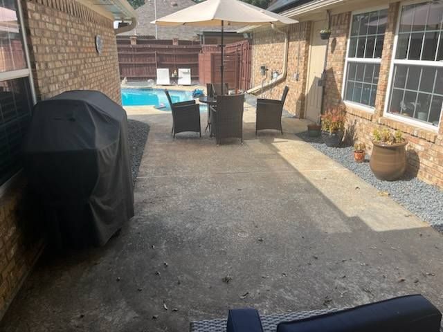 Back patio with grill, table and chairs beside a house and swimming pool.