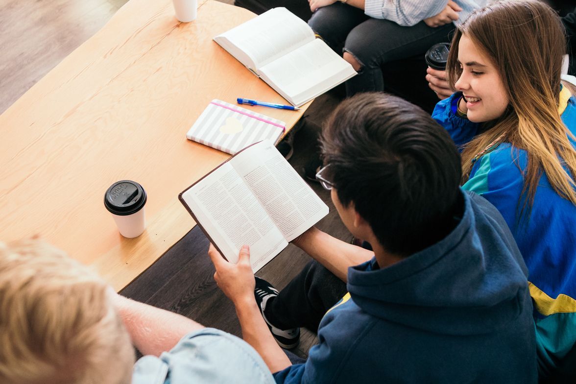 Group of students studying together at a table, one looking at a notebook, others with drinks and books.