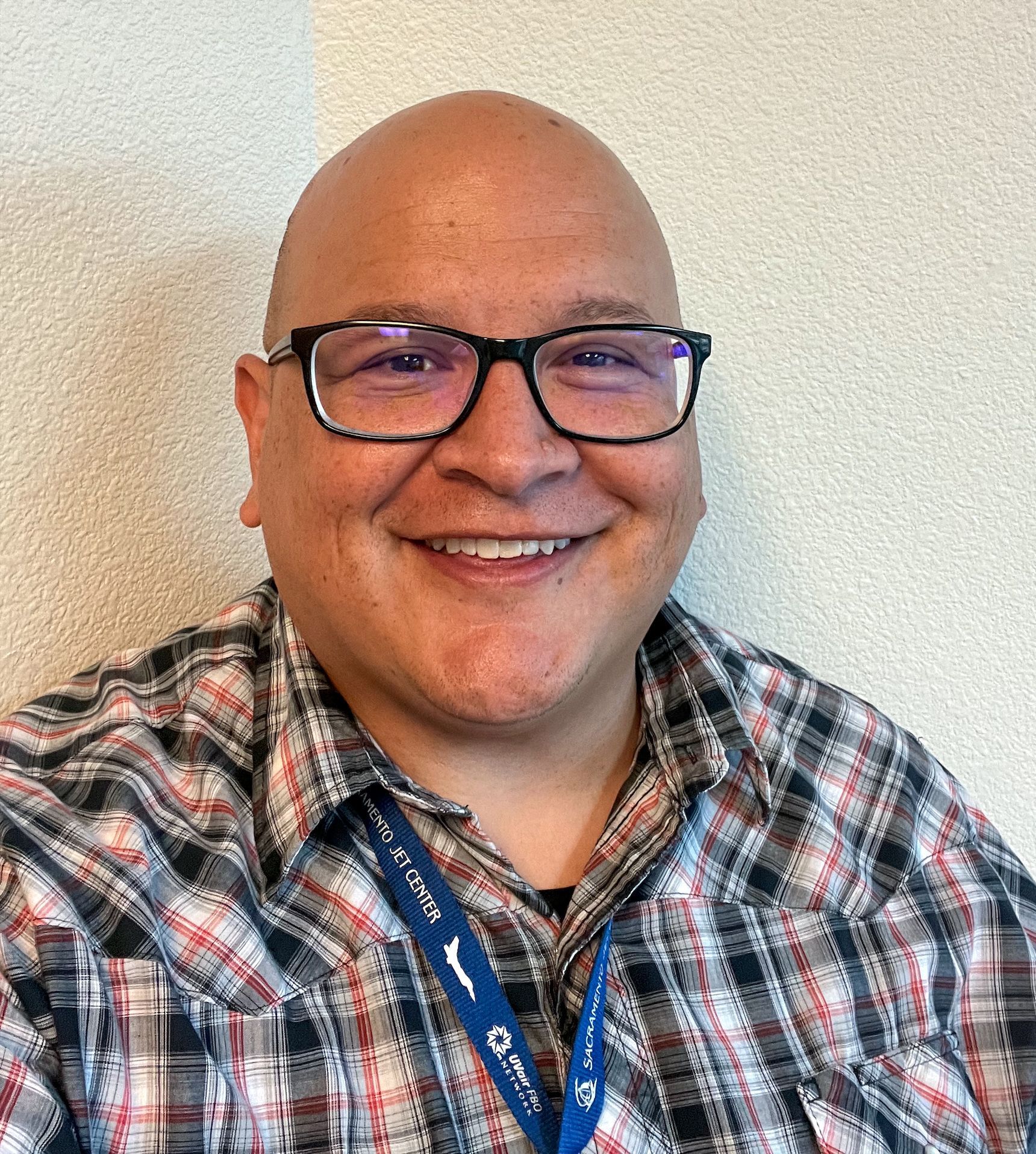 Smiling person in glasses and plaid shirt with a blue lanyard against a light wall