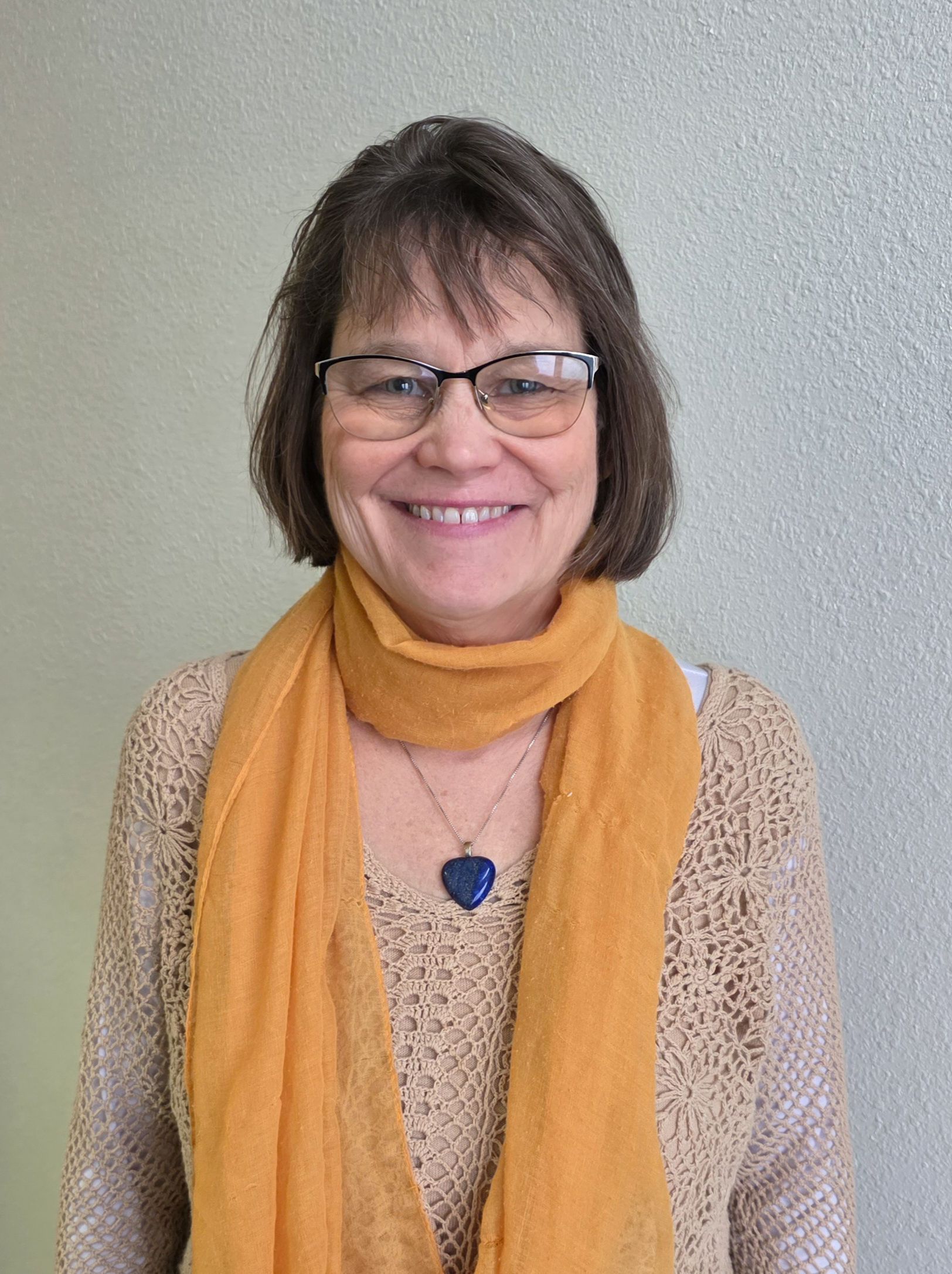 Smiling person wearing glasses, a mustard scarf, and a blue pendant against a gray wall.
