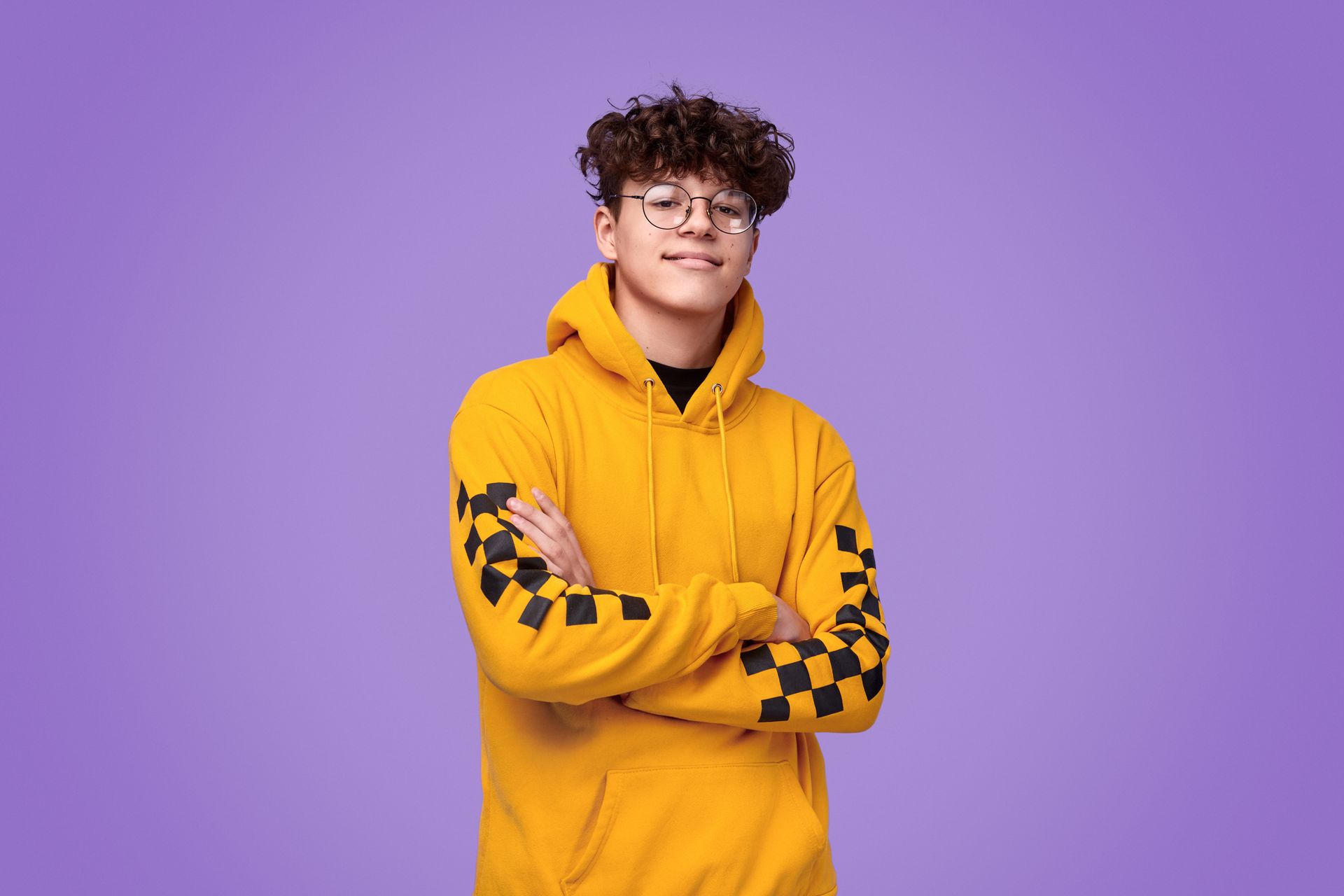 A young man wearing a yellow hoodie and glasses is standing with his arms crossed.