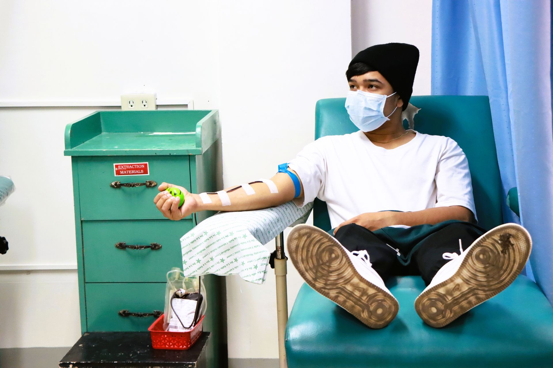 Person wearing a mask donates blood in a clinic setting, holding a stress ball, with an IV in arm.