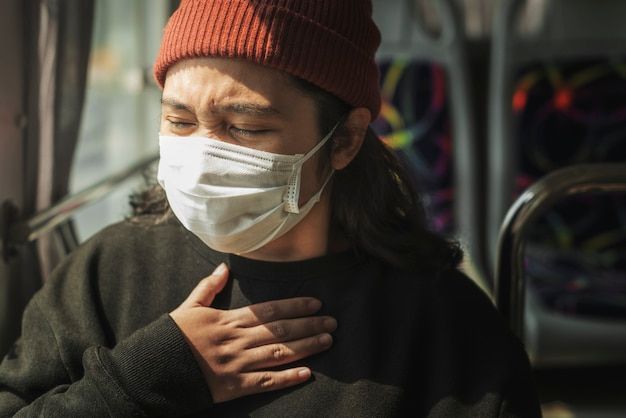 Person wearing a mask on a bus, clutching chest, eyes closed, wearing a knit cap.