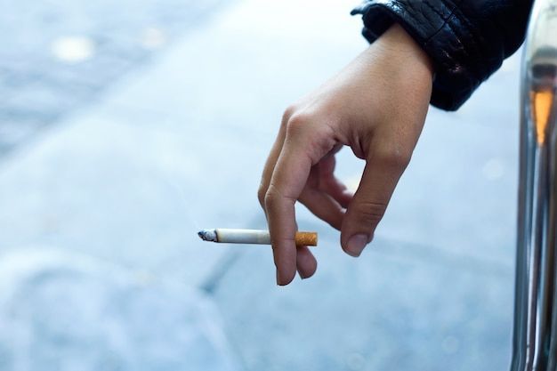 Hand holding a lit cigarette, indoors near a metal railing, blurred background.