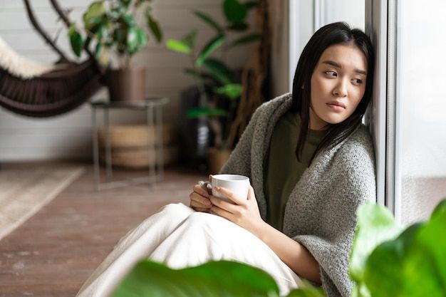 Woman sits by window, wrapped in blanket, holding mug, looking wistful.