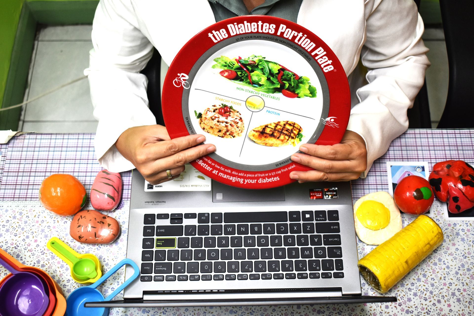 Person holding a red diabetes portion plate over a laptop surrounded by colorful toy food.