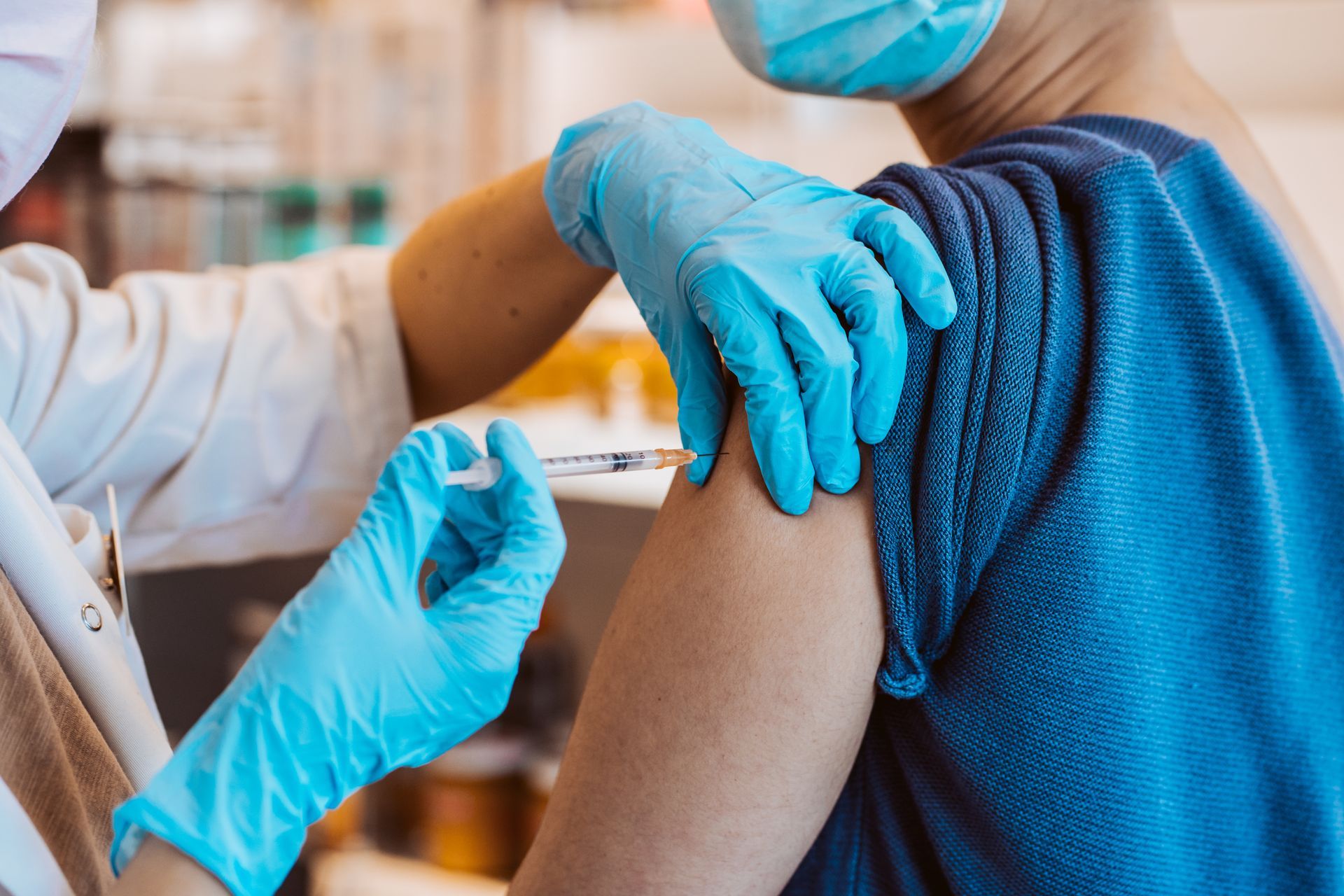 Person receiving a vaccine injection in the upper arm, indoors. Healthcare worker wearing blue gloves and mask.