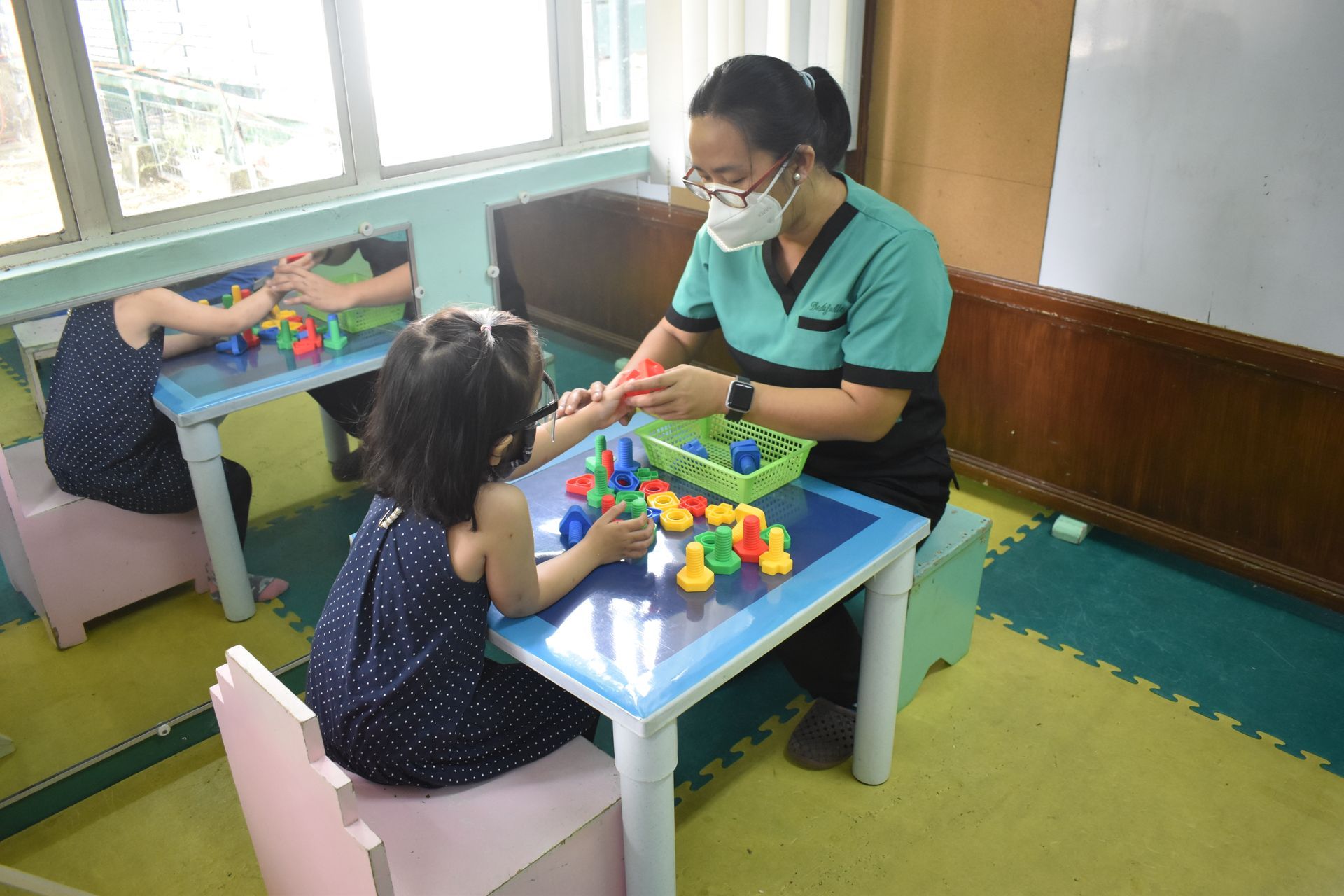 A person in a mask assists a child with colorful building blocks at a table in a brightly lit room.