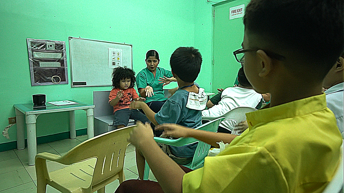 Children in a room with a medical professional. One child sits on a chair. Others are seated in chairs.