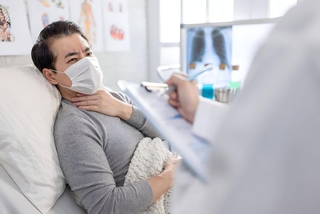 Man in bed wearing mask, holding throat, examined by doctor with clipboard.