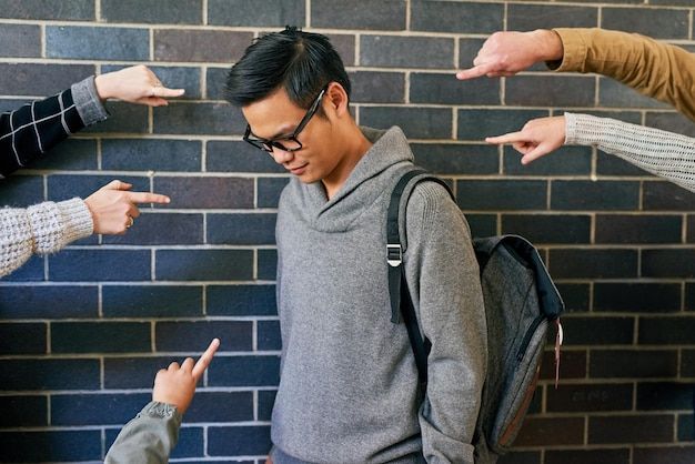 Man with glasses, backpack, and downcast expression, being pointed at by multiple hands.