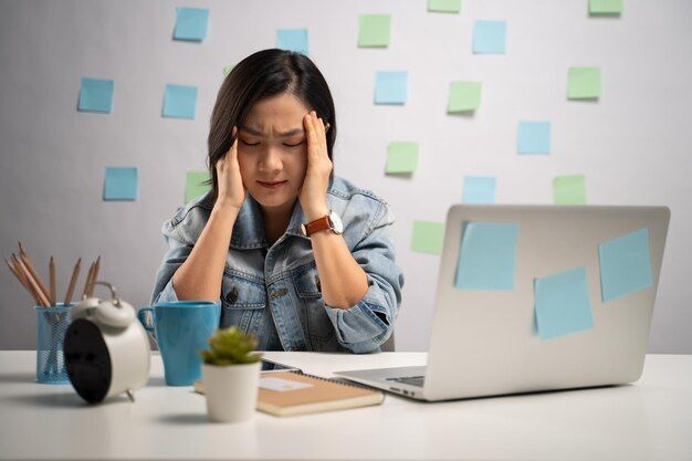 Woman with hands on head, stressed at desk with laptop, sticky notes, clock and plant.